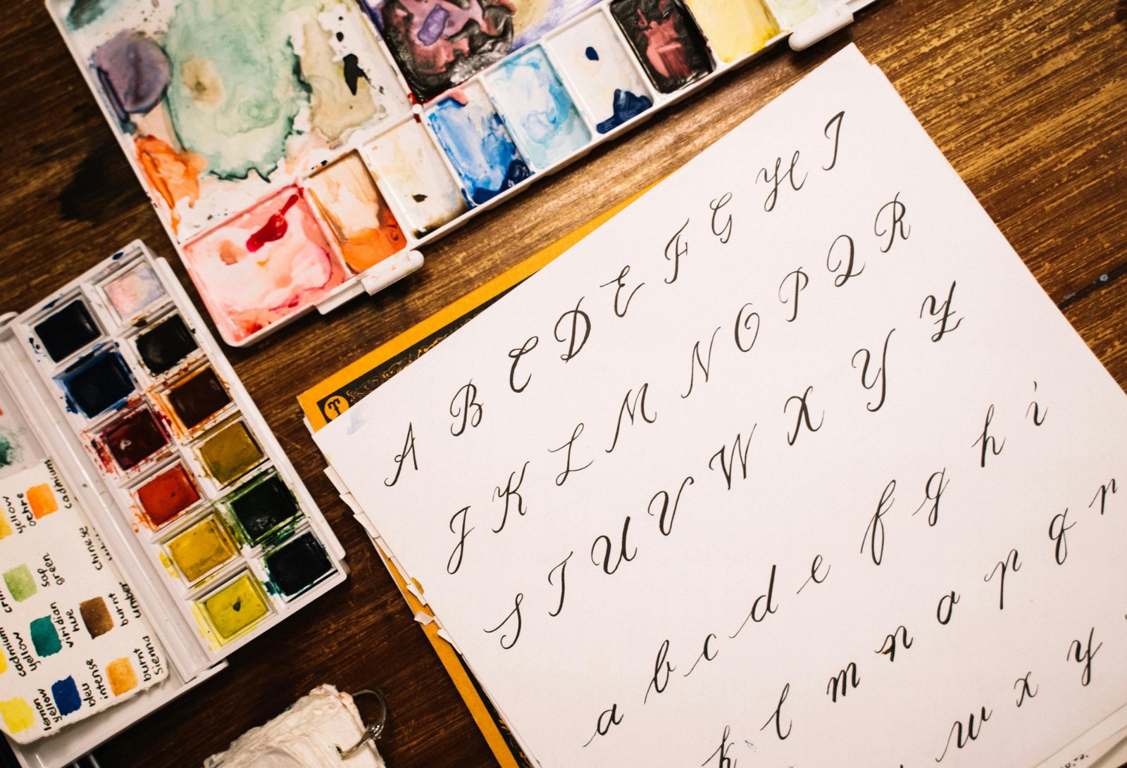 Brush lettering alphabet practice sheet next to two watercolor palettes